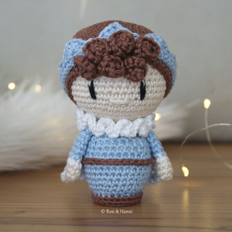 Gehäkelte Amigurumi-Puppe mit hellem Gesicht, blauem Kleid, Rüschenkragen und braunen Locken