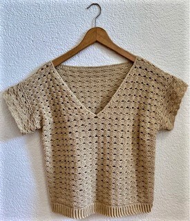 Beigefarbenes gehäkeltes Damen-Shirt mit V-Ausschnitt und kurzen Ärmeln an Holzbügel vor heller Wand.