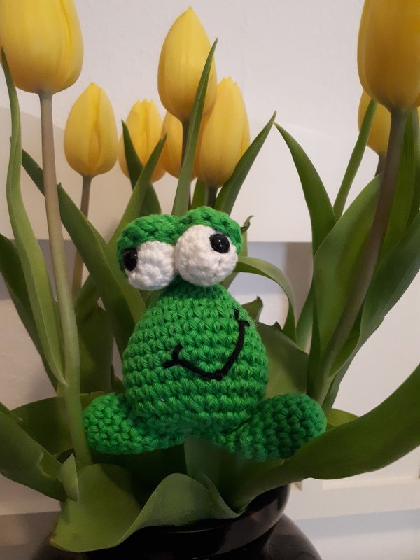 Gehäkelter grüner Frosch mit großen weißen Augen und einem Lächeln zwischen gelben Tulpen und grünen Blättern.