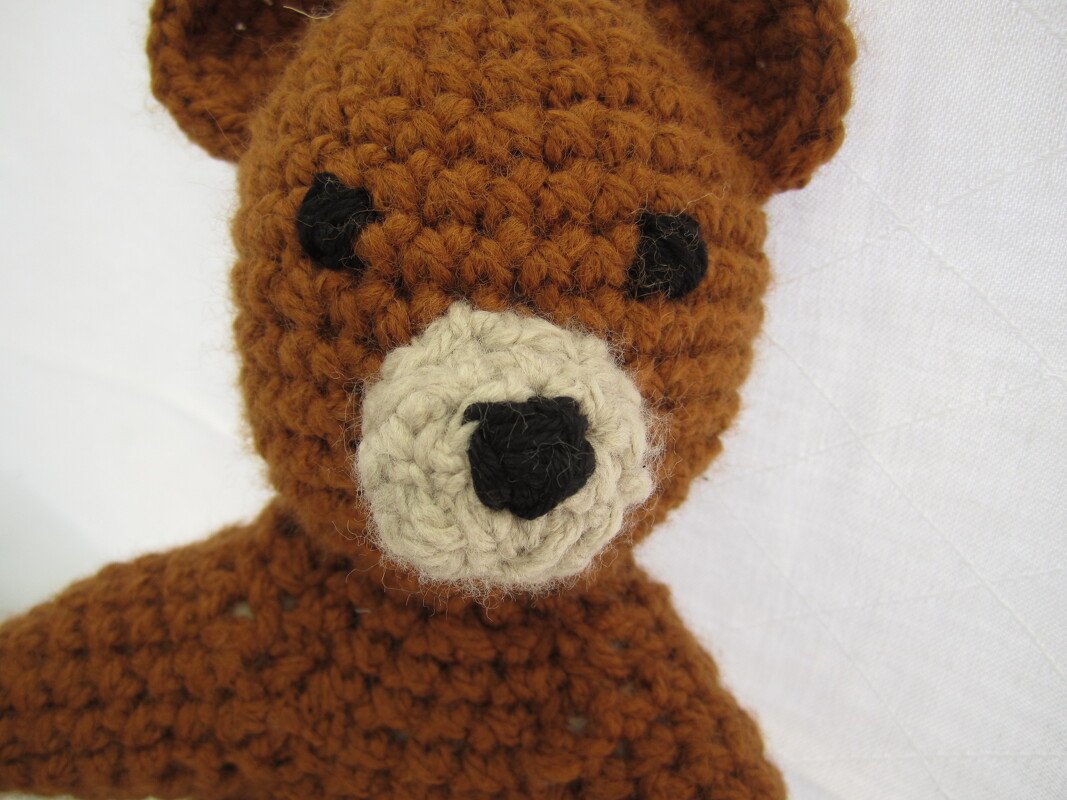 Gehäkelter brauner Teddybär mit schwarzen Augen und beiger Schnauze vor hellem Hintergrund.