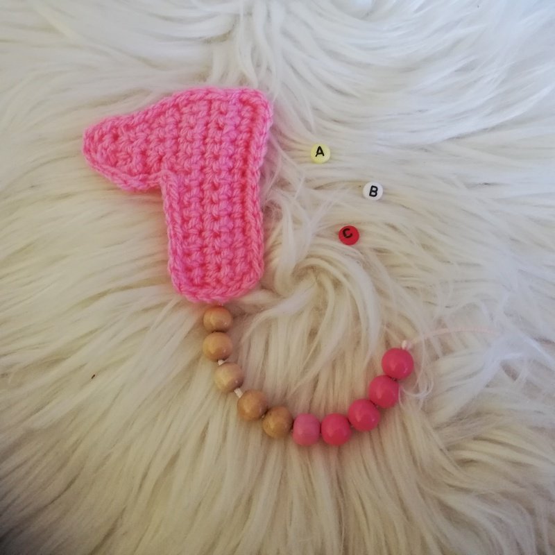 Gehäkelte rosa Zahl 1 mit natur- und rosafarbenen Holzperlen auf weißem Plüsch