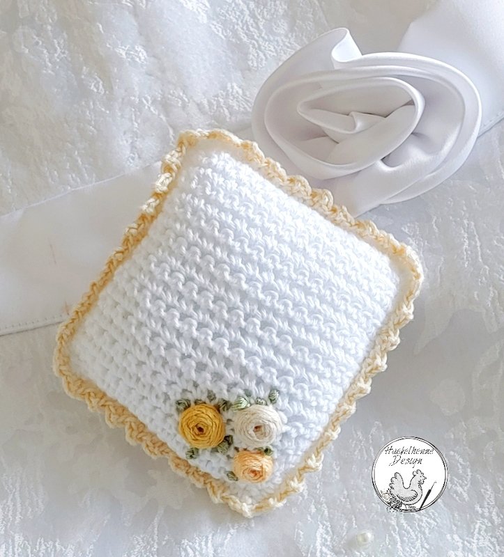Weißes gehäkeltes Ringkissen mit cremefarbenem Rüschenrand und drei kleinen Rosenapplikationen in Gelb und Creme.
