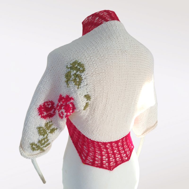 Kurz gestrickter cremefarbener Damen‑Bolero mit rotem Netzeinsatz im Rücken und Rosenintarsia am Ärmel auf einer Schaufensterpuppe