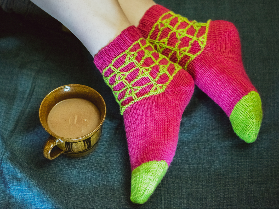 Füße in rosa-grünen gestrickten Socken mit netzartigem Muster neben einer Tasse auf dunklem Stoff