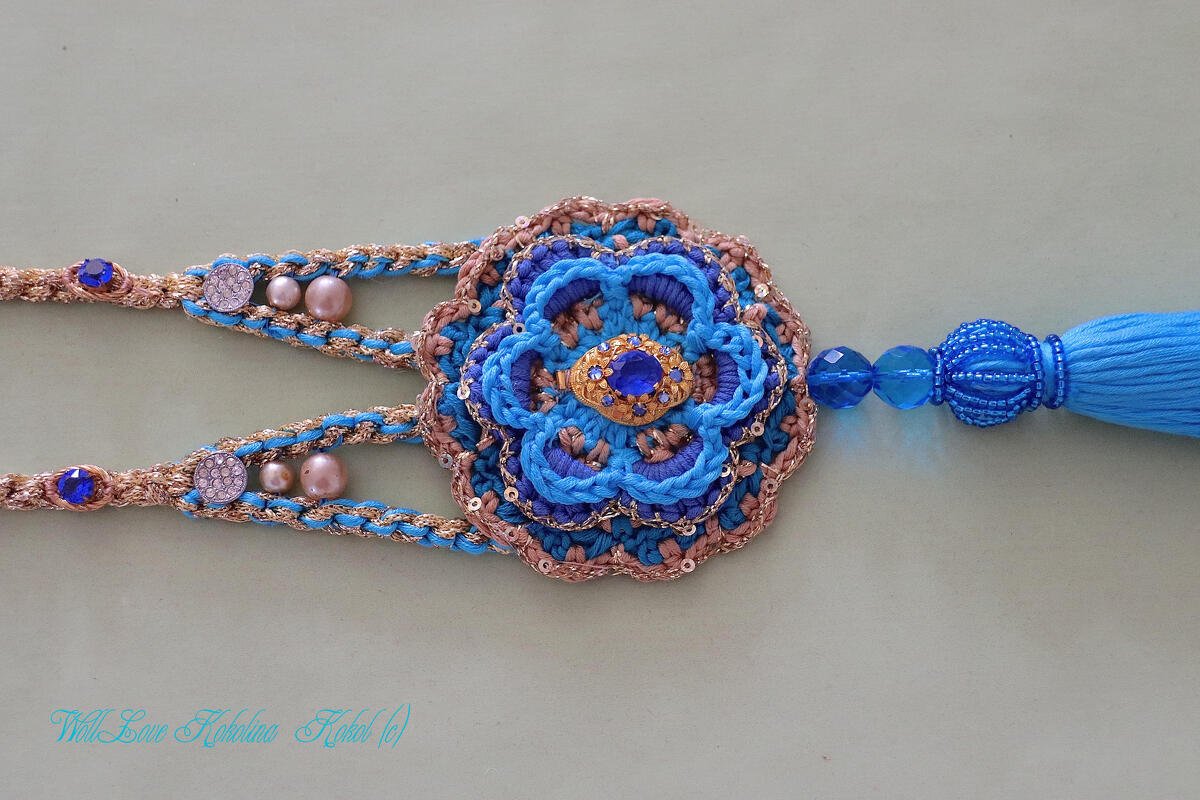 Nahaufnahme eines handgehäkelten Accessoires in Blau und Gold mit zentraler Blüte, blauem Schmuckstein, Perlen, Pailletten und einer blauen Quaste.