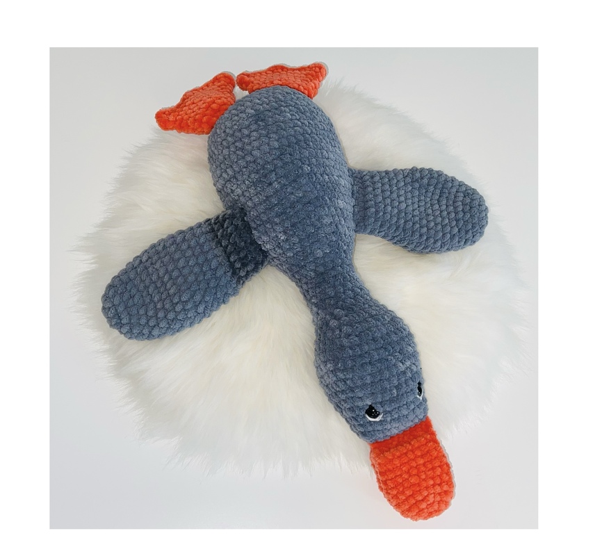 Gehäkelte graublaue Amigurumi-Gans mit orangefarbenem Schnabel auf weißem Felluntergrund