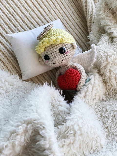 crocheted amigurumi angel doll with yellow hair, halo, white wings holding a red heart on a pillow and fluffy blanket