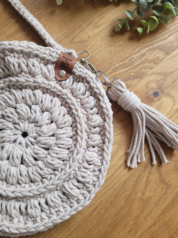 Runde gehäkelte Umhängetasche in Beige mit Reliefmuster, Trageriemen und Quaste auf Holz.