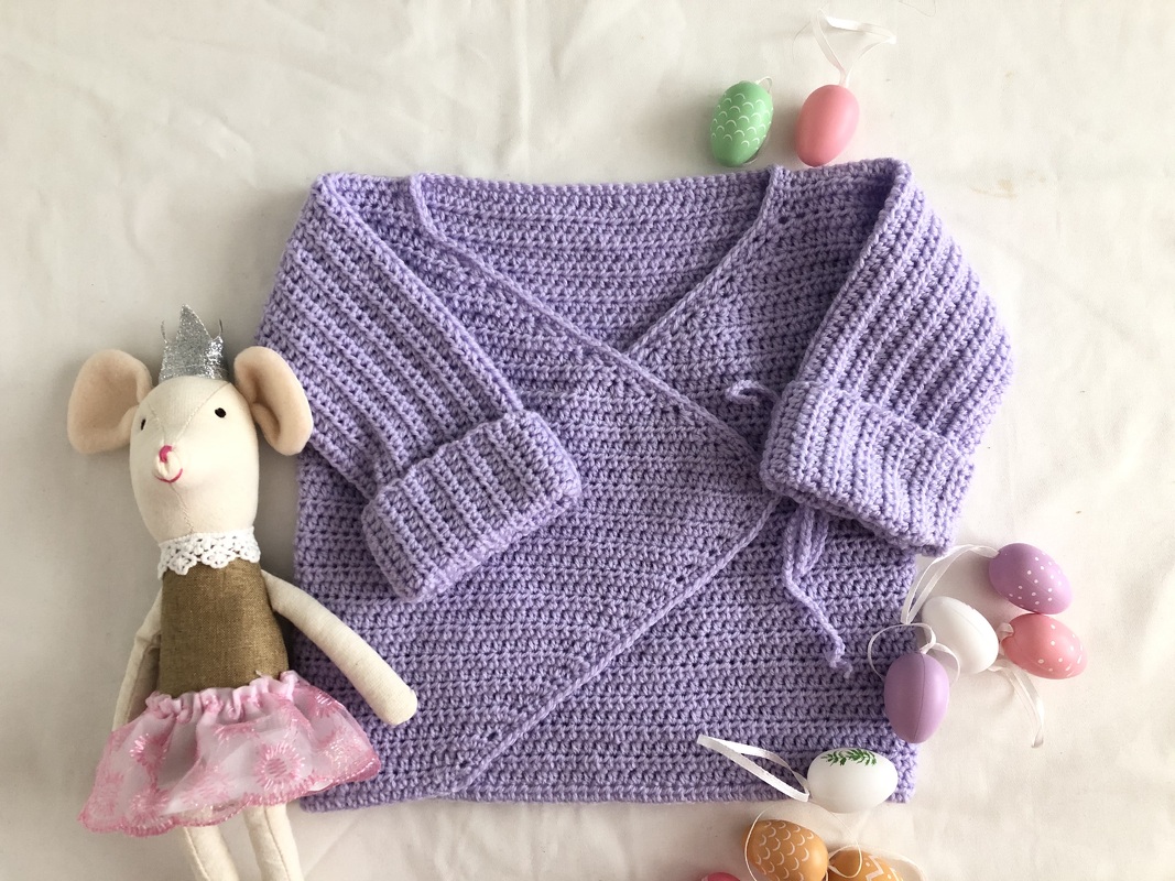 Light purple crocheted baby wrap jacket with long sleeves, ribbed cuffs and tie closure next to a mouse toy and pastel eggs.