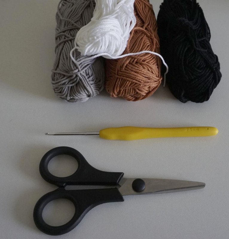 Four yarn skeins in gray, white, brown and black with a yellow-handled crochet hook and small craft scissors on a light surface