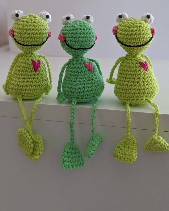 Drei gehäkelte Amigurumi-Frösche in Hell- und Dunkelgrün mit aufgenähten Herzen, sitzend an einer Kante