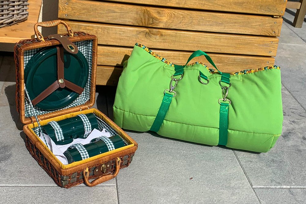 Gepolsterte grüne Decke mit stabilen Gurten, Karabinern und D‑Ringen neben geöffnetem Picknickkorb mit Geschirr