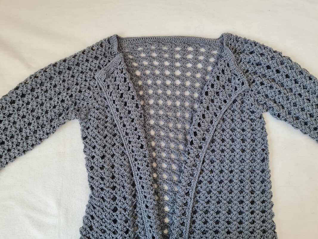 Graue gehäkelte Damenjacke mit langen Ärmeln und durchgehendem Lochmuster, offen ausgebreitet auf hellem Untergrund.