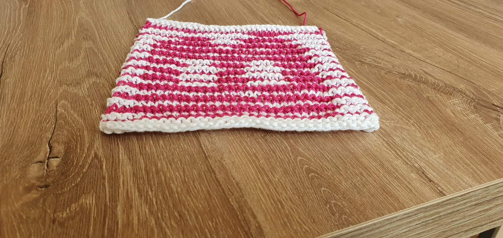 Gestrickte Maschenprobe in Pink und Weiß mit Herzmotiv auf Holzuntergrund