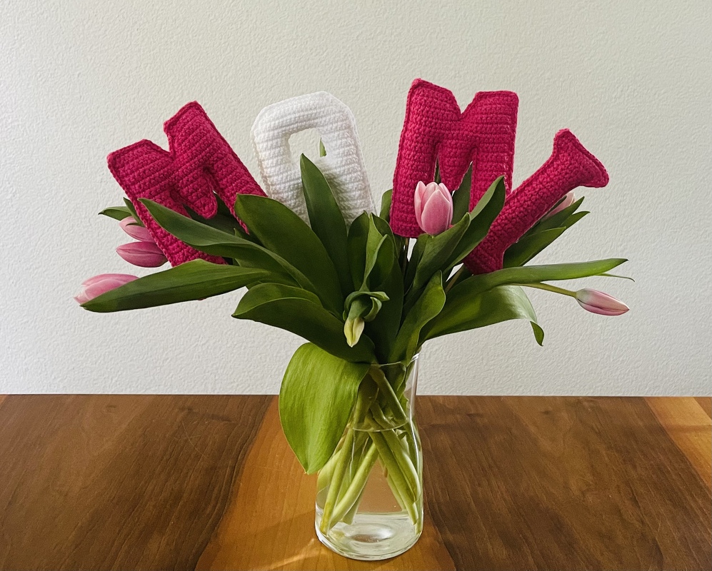Gehäkelte MOM-Buchstaben in Pink und Weiß zwischen rosa Tulpen in einer Glasvase auf einem Holztisch