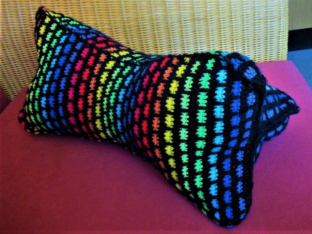 Längliches gehäkeltes Kissen mit buntem Regenbogen-Gittermuster auf schwarzem Grund, auf magentafarbenem Untergrund vor Korbgeflecht.