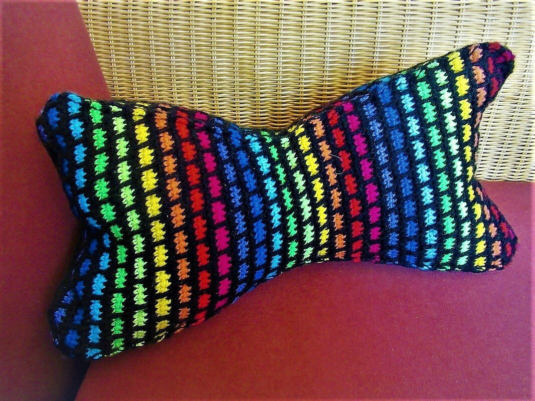 Gehäkeltes Knochenkissen mit bunten Regenbogenstreifen auf schwarzem Grund, auf rotem Sofa vor Rattangeflecht