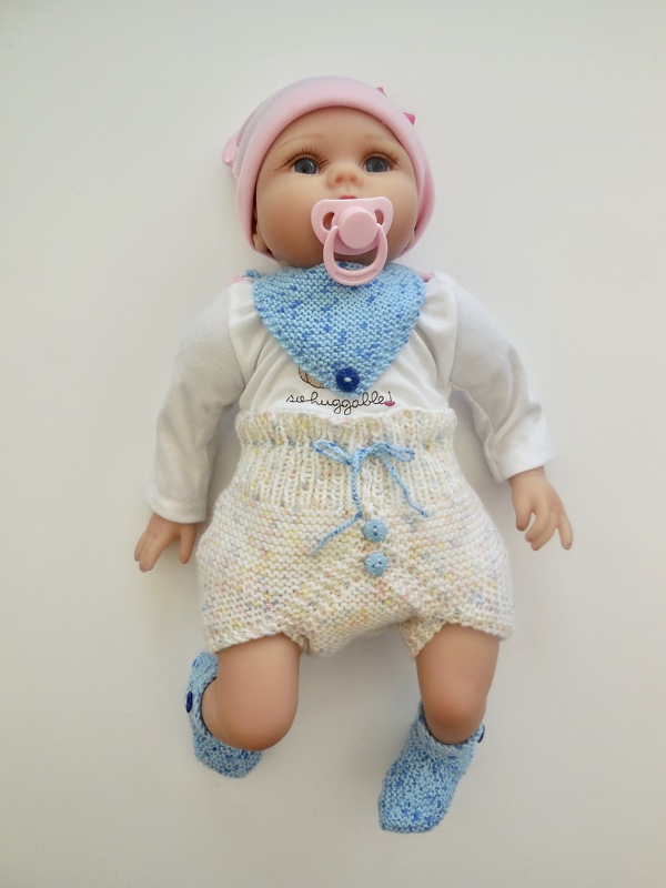 Babypuppe in gestricktem Baby‑Set: dreieckiges Lätzchen, kurze Hose mit Kordelzug und Zierknöpfen sowie passende Söckchen in Pastellfarben.