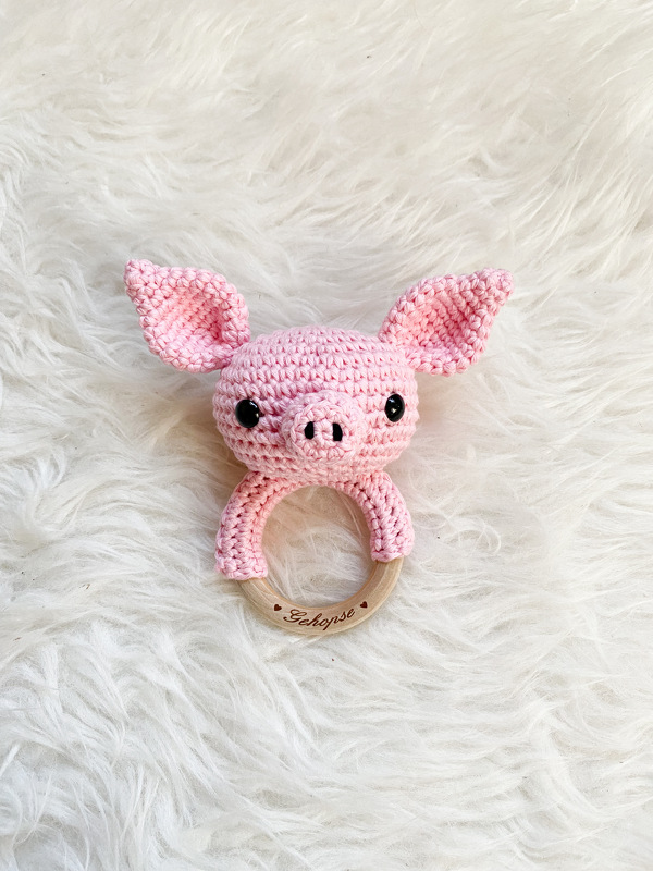 Rosa gehäkelter Schweinekopf, befestigt an einem hellen Holzring, liegend auf einer plüschigen weißen Unterlage.