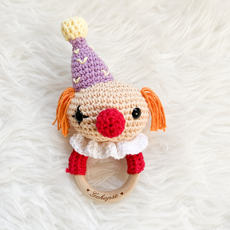 Gehäkelte Clown-Rassel mit lila Hütchen, orangefarbenen Zöpfen und roter Nase an einem Holzring mit Gravur 'Gehopse', auf weißem Kunstfell.