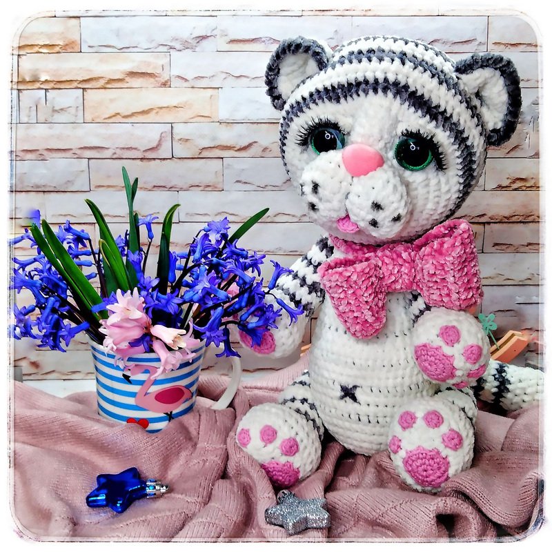 White and gray striped crocheted amigurumi cat with pink bow sitting on a pink blanket next to a mug of hyacinth flowers.