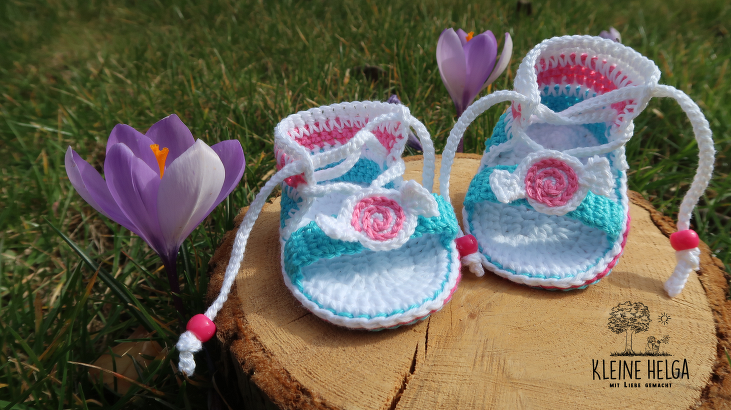 Gehäkelte Babysandalen in Weiß, Türkis und Rosa mit Blumenapplikation und Kordel auf Holzscheibe vor Gras und Krokussen