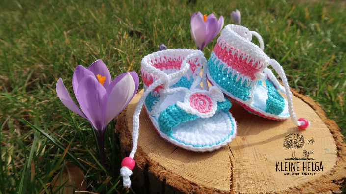 Gehäkelte Babysandalen in Pastellfarben mit Blüte auf Holzscheibe im Gras
