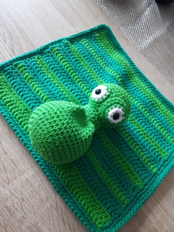 Gehäkeltes grünes Amigurumi-Drachenbaby mit großen weißen Augen auf grün gestreifter Häkelunterlage.