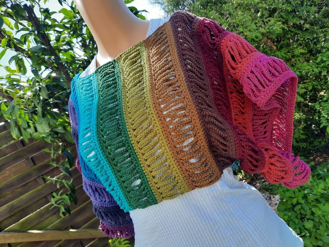 Gehäkelter Poncho in Regenbogenfarben mit offenem Muster auf Schaufensterpuppe vor Gartenpflanzen.