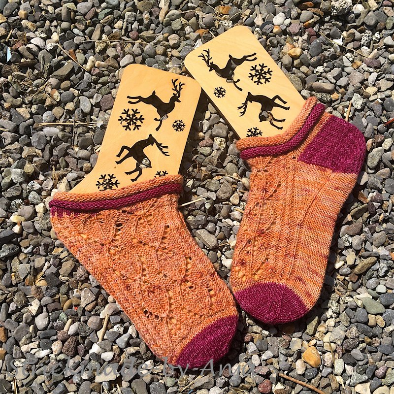 Zwei kurze gestrickte Damensocken in Orange und Dunkelrot mit filigranem Ajourmuster auf Holzsockenbrettern mit Rentier- und Schneeflockenmotiven, liegend auf Kieselsteinen.