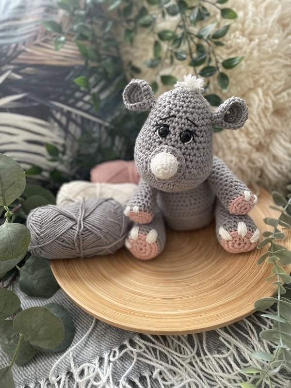 Gehäkeltes graues Amigurumi‑Nilpferd mit rosa Pfoten auf Holzschale neben Wollknäuel