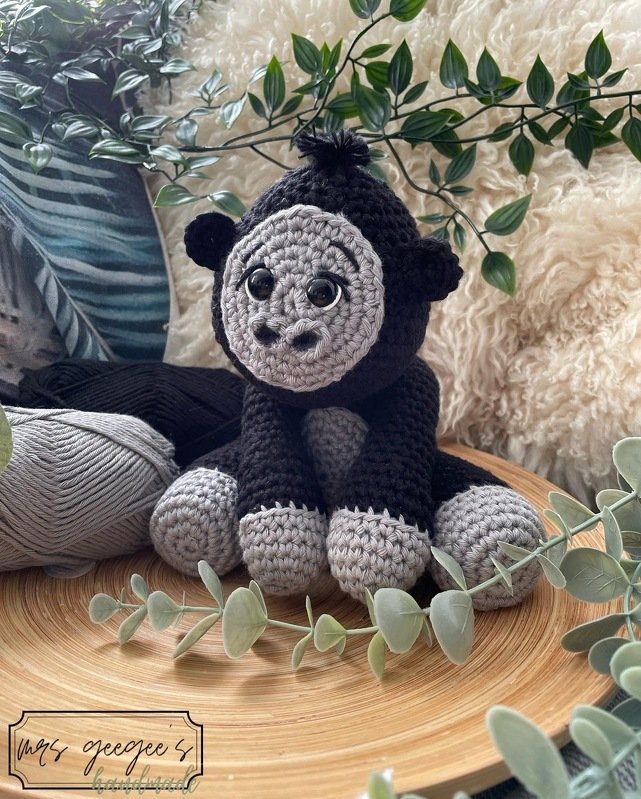 Gehäkelter Amigurumi-Gorilla in Schwarz und Grau mit großen glänzenden Augen auf Holzscheibe, umgeben von Garn und grünen Blättern