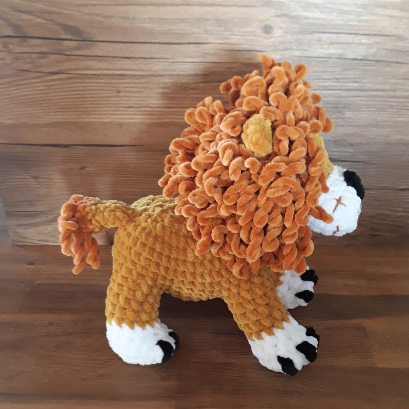 Gehäkelter Amigurumi-Löwe mit braun-orangefarbenem Körper, flauschiger orangefarbener Mähne und weißen Pfoten auf Holzuntergrund.