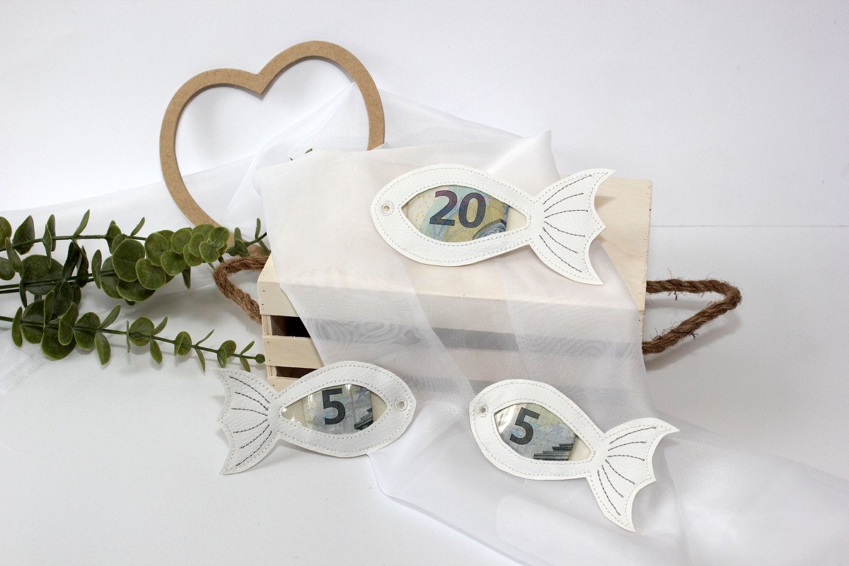 Weiße Fisch‑Anhänger mit Sichtfenster und Geldscheinen auf Holzkiste, Herzdekoration und Grünzweigen