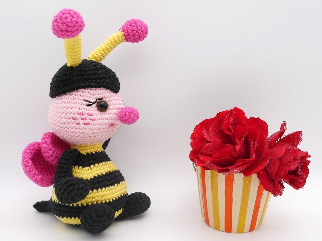 Gehäkelte Amigurumi-Biene in Schwarz-Gelb mit rosa Kopf und Flügeln neben roten Rosen im gestreiften Becher