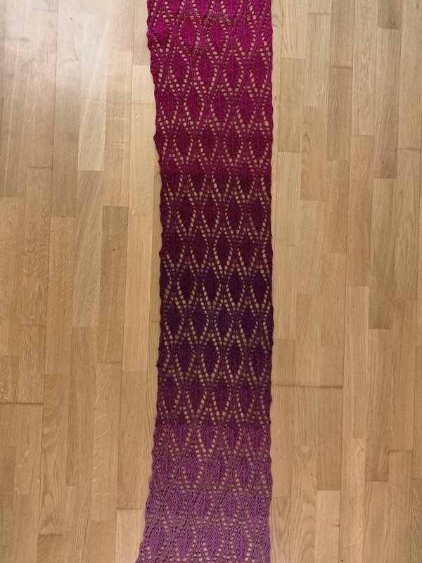 Long crocheted lace scarf with diamond openwork and magenta-to-lilac ombré laid flat on a wood floor