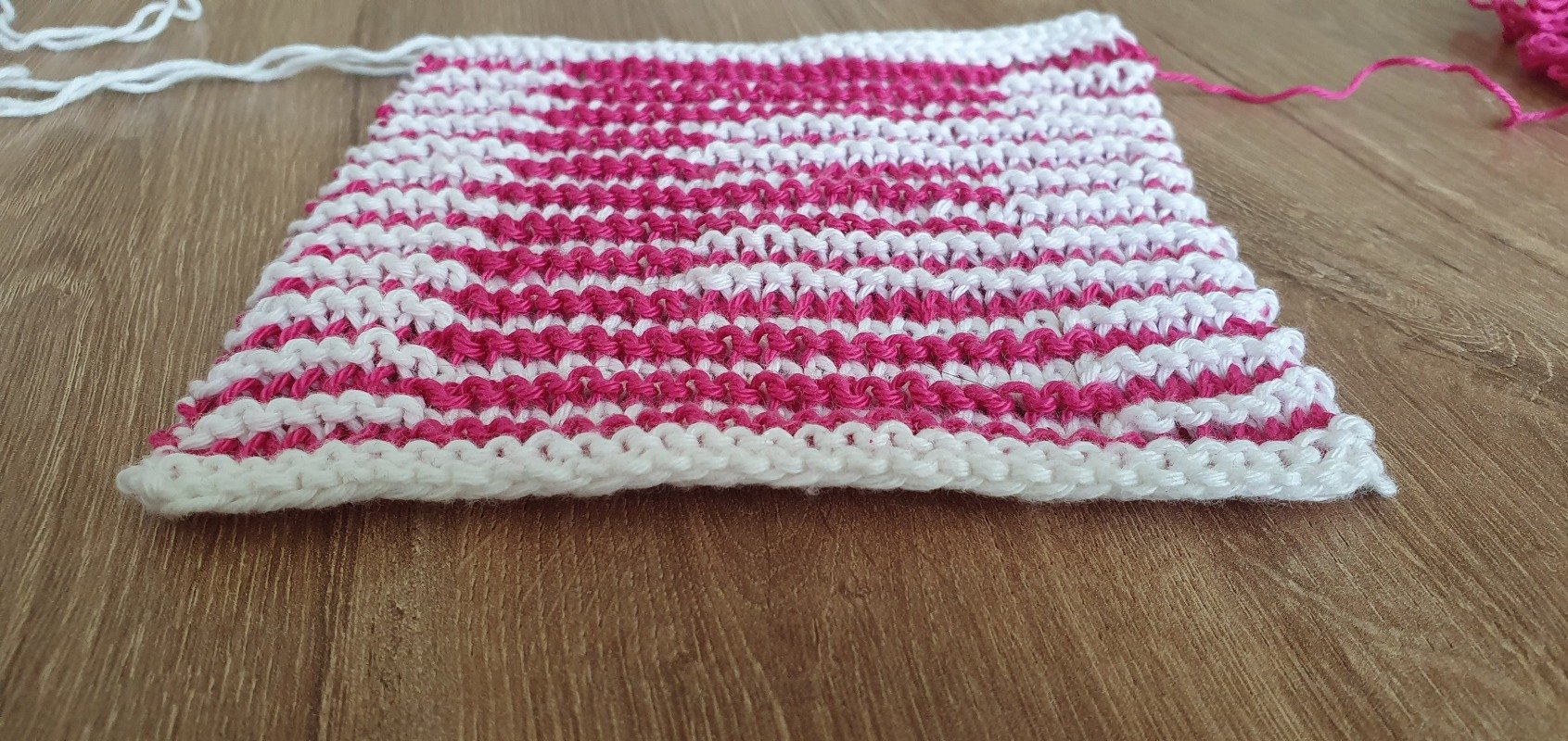 Zweifarbige Strickprobe mit pinken und weißen Reihen auf Holz, losen Garnenden