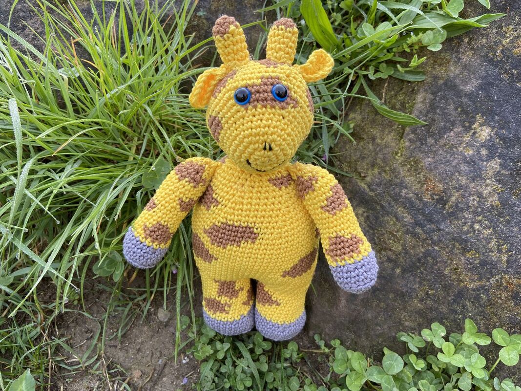 Gehäkelte Amigurumi-Giraffe in Gelb mit braunen Flecken, blauen Augen und grauen Hufen, im Gras neben einem Stein.