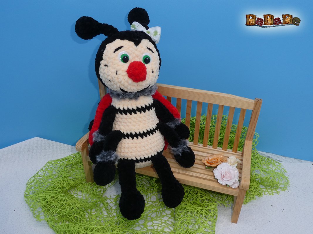Handgehäkelter Marienkäfer-Amigurumi mit roten Flügeln, schwarzem Körper und grünen Kunststoffaugen sitzt auf einer kleinen Holzbank neben zwei Papierblumen.