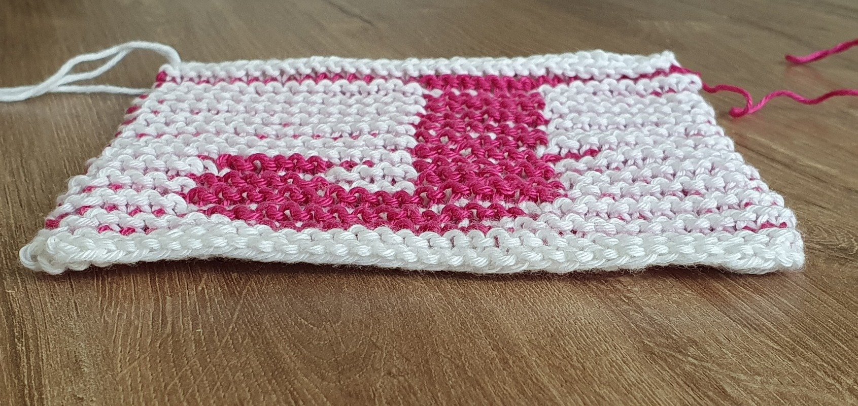Nahaufnahme einer gestrickten Probe in Weiß mit pinkfarbenem Buchstabenmuster auf Holz.
