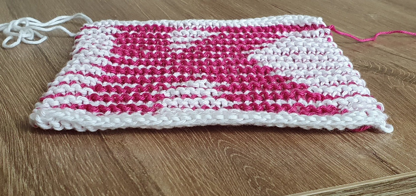 Gestrickte Maschenprobe in Weiß und Pink mit sichtbarer Seitenkante auf Holzboden