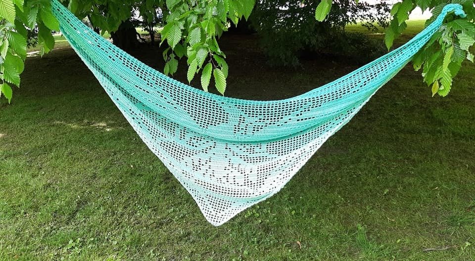 Gehäkeltes Dreieckstuch in Mintgrün und Weiß mit feinem Spitzenmuster, zwischen Baumzweigen aufgehängt über einer Wiese.