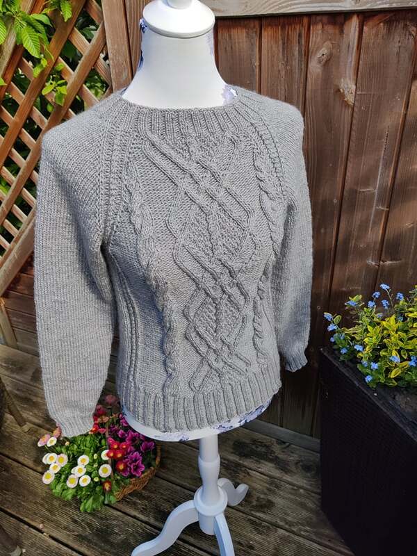 Gestrickter grauer Pullover für Damen mit Zopfmuster auf einer Schneiderpuppe vor Holzzaun