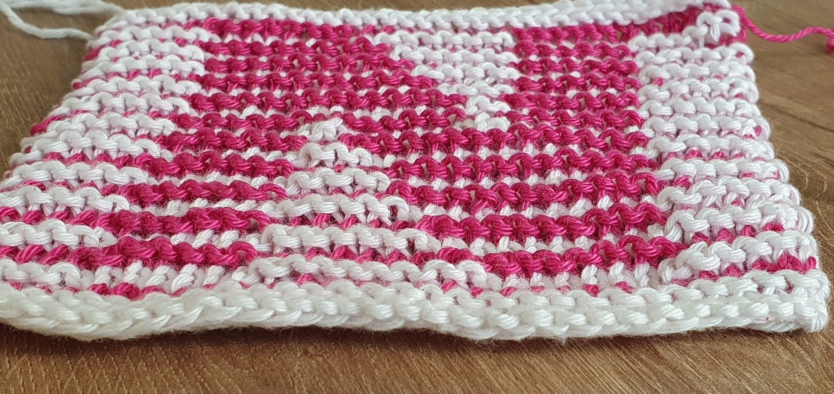 Gestrickte Musterprobe in Pink und Weiß mit wiederholtem Herz- und Rautenmuster auf Holzuntergrund.