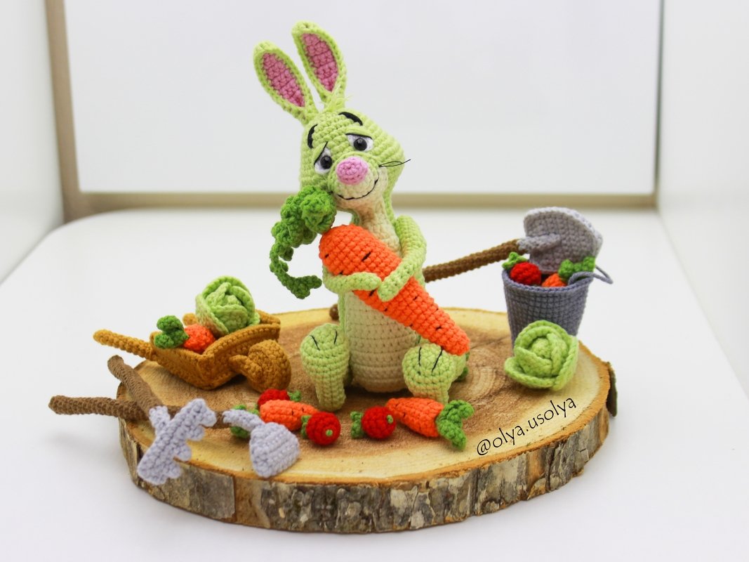 Green crocheted amigurumi rabbit holding an orange carrot with crocheted vegetables on a wood-slice base