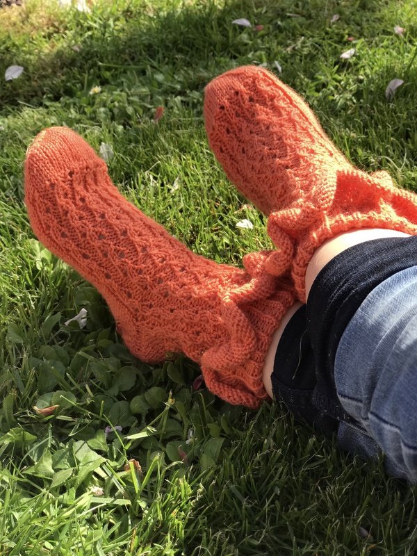 Orangefarbene handgestrickte Stricksocken mit durchbrochenem Spitzenmuster und Rüschenbündchen auf grünem Gras.