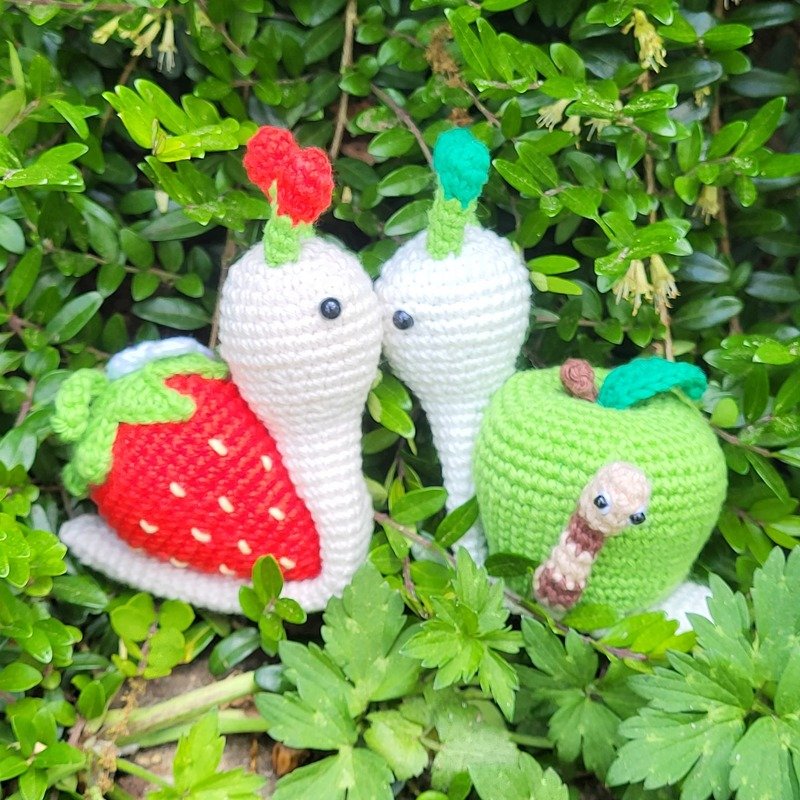 Zwei gehäkelte Amigurumi‑Schnecken, eine mit Erdbeer‑Panzer, neben einem grünen Apfel mit kleinem Wurm im Blattgrün.