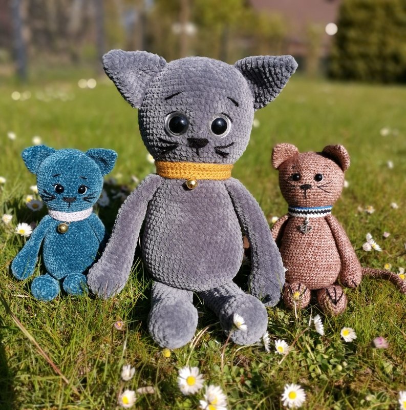 Drei gehäkelte Amigurumi-Katzen in Blau, Grau und Braun sitzen auf einer Wiese