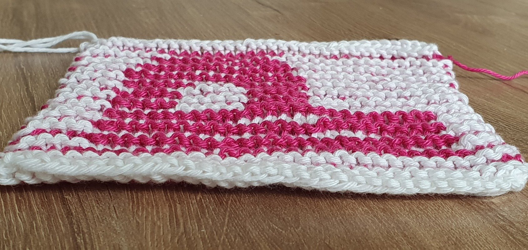 Flache Nahaufnahme einer Strickprobe mit pinkem Motiv auf weißem Grund, sichtbare Maschenkanten und Fadenenden