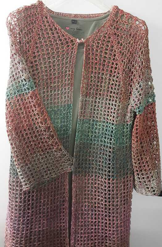 Lange offene Häkeljacke in Rosé, Grün und Violett mit lockerem Netzmuster, über einem olivgrünen Shirt an einem Kleiderbügel hängend.